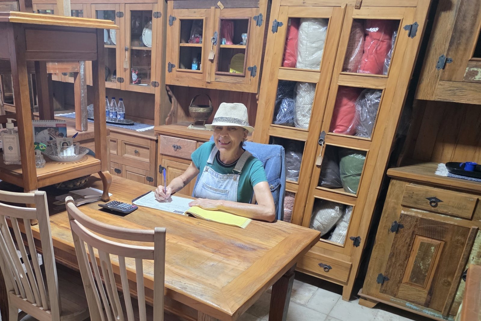 Dona Leila Maria sentada à mesa de atendimento no meio da loja, escrevendo um pedido, cercada de cristaleiras e aparadores de madeira